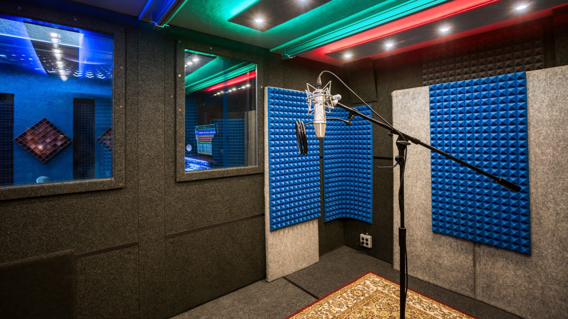 Isolated vocal recording booth with studio microphone, acoustic foam treatment, control room viewing window, and professional lighting system