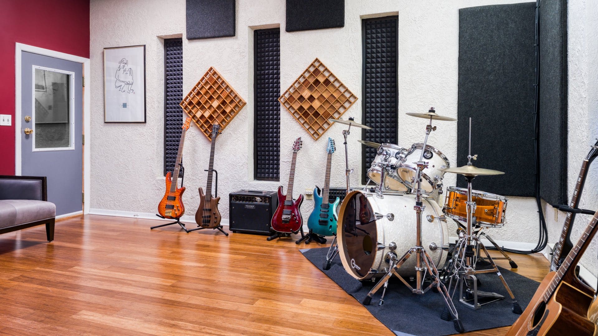 Spacious recording studio with drum kit, multiple electric guitars, bass guitars, amplifiers, acoustic treatment panels, and professional lighting on hardwood floors