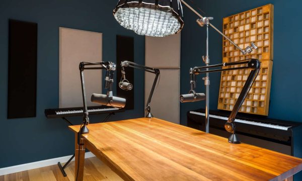 Professional podcast studio with wooden desk, broadcast microphones on boom arms, acoustic panels, reflector umbrella, and keyboard setup
